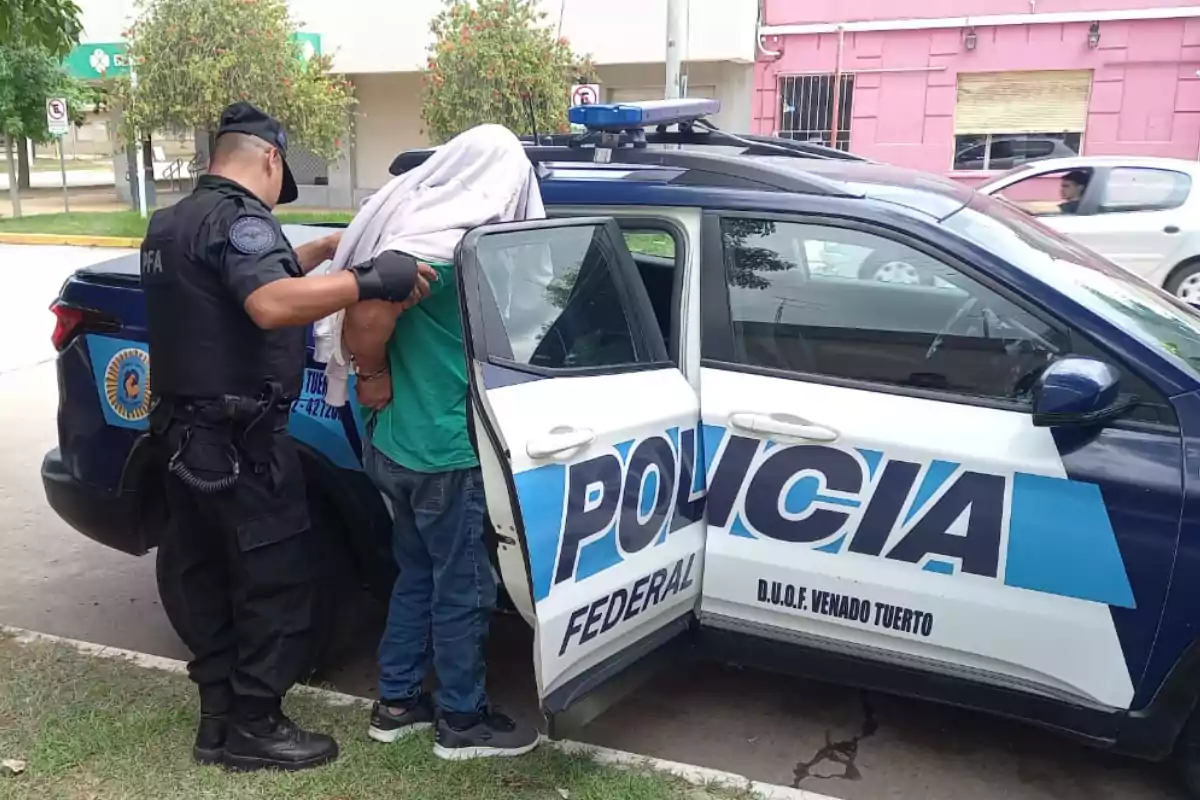 Un oficial de policía escolta a una persona hacia un vehículo policial con la puerta trasera abierta.