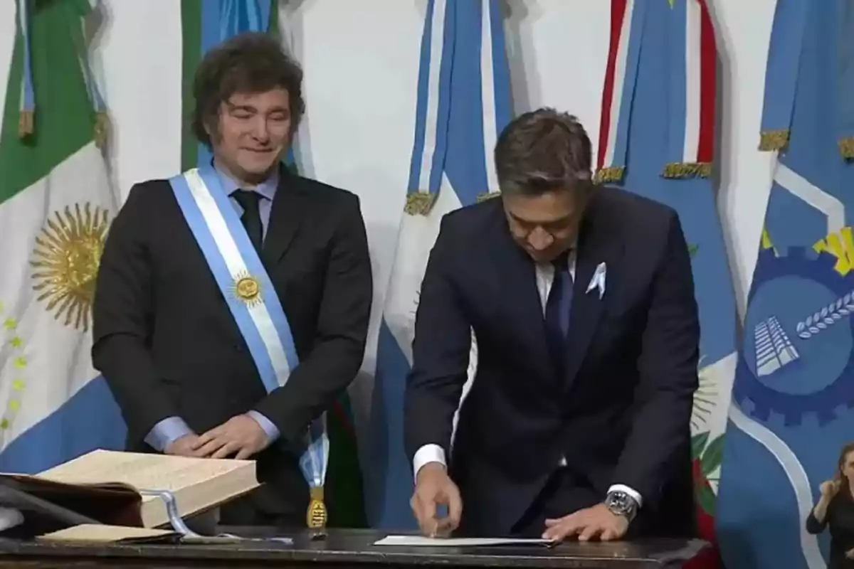 Two men in formal suits are at an official event with Argentine flags in the background; one of them is wearing a presidential sash and the other is signing a document on a table.