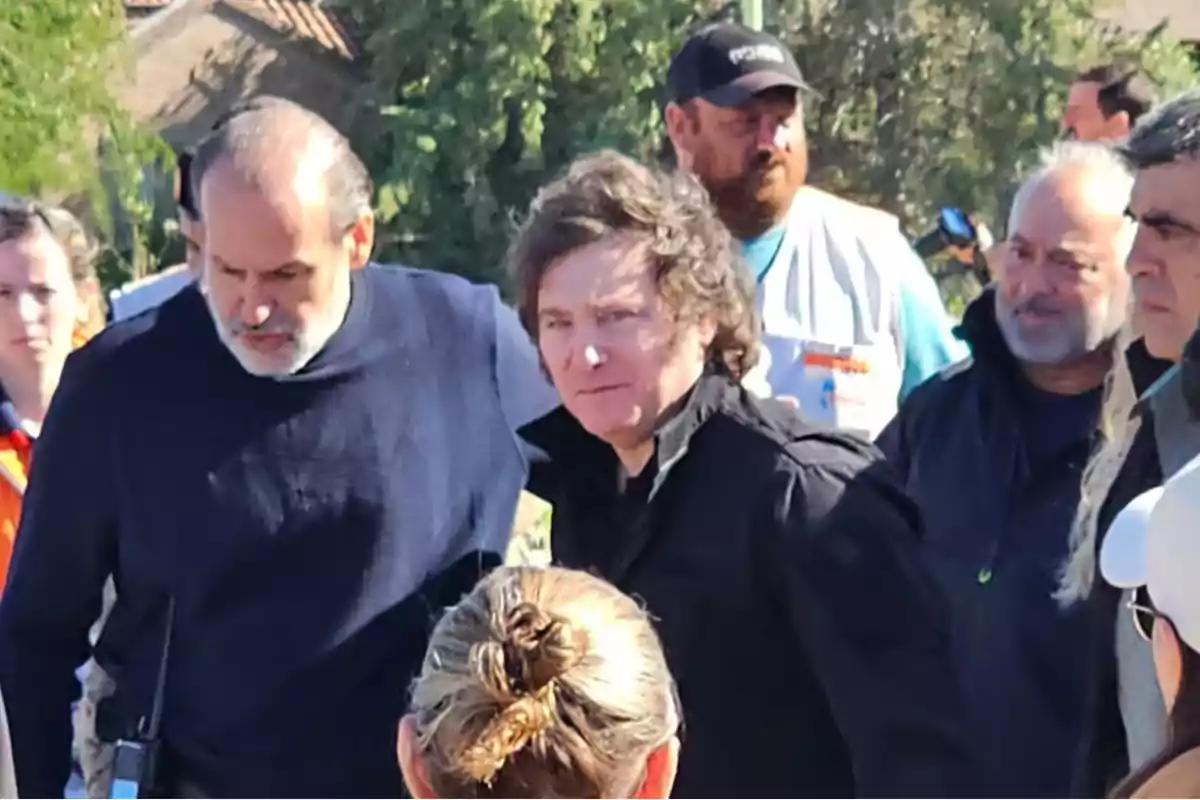 Un grupo de personas está reunido al aire libre, algunas de ellas llevan ropa de trabajo y chalecos, mientras que otras parecen estar conversando o prestando atención a algo fuera de la imagen.