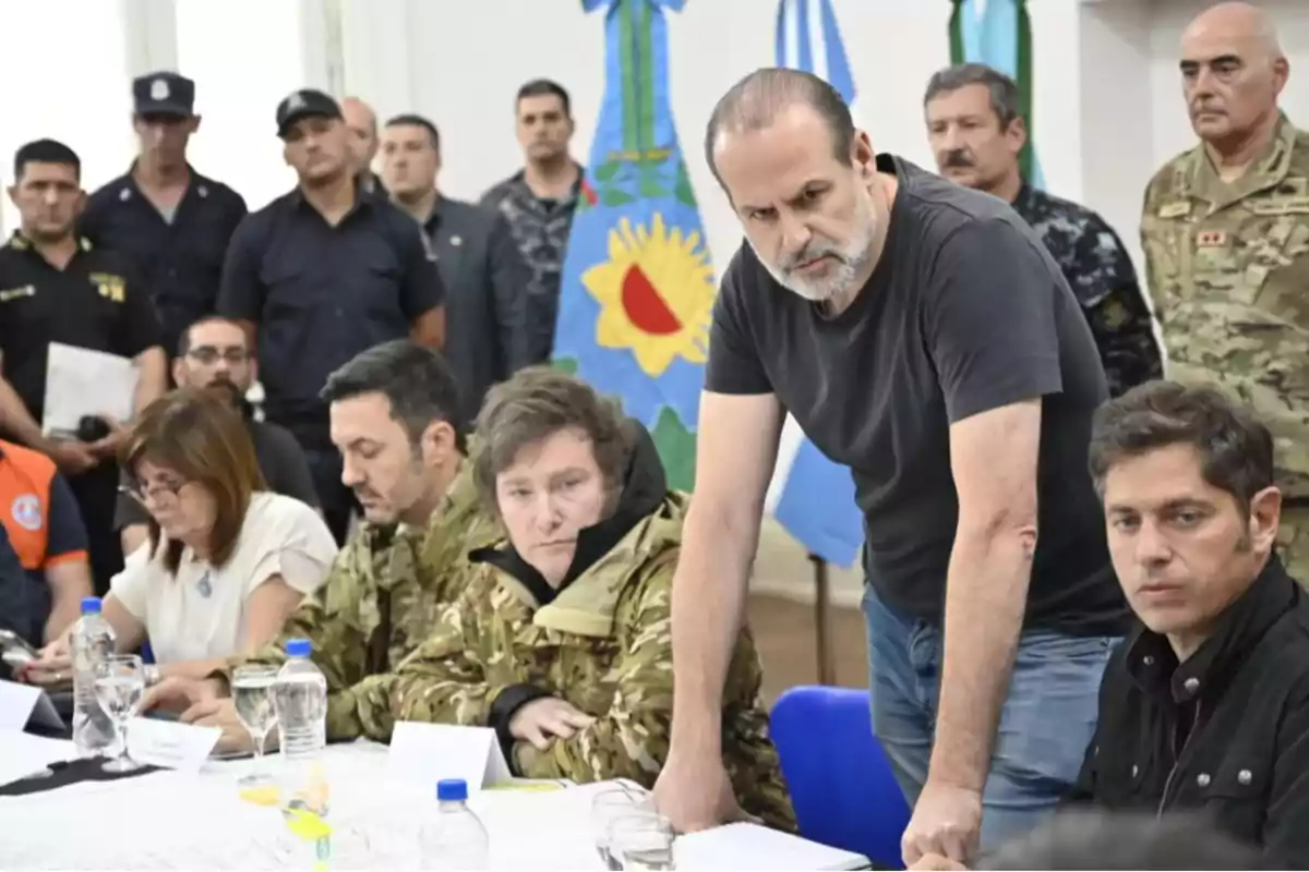 Un grupo de personas, algunas en uniforme militar, se encuentra reunido alrededor de una mesa con documentos y botellas de agua, mientras un hombre de pie parece estar hablando o dirigiendo la reunión.