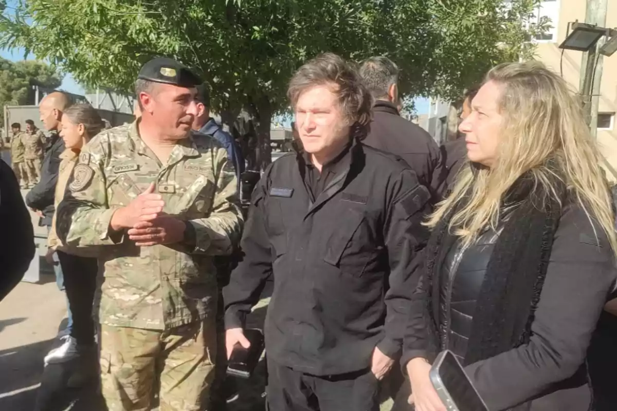 Personas vestidas con uniformes militares y ropa oscura conversan al aire libre en un entorno con árboles.