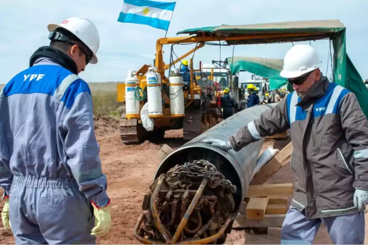 YPF workers with helmets and uniforms inspect a large metal pipe at a construction site, with heavy machinery and an Argentine flag waving in the background.