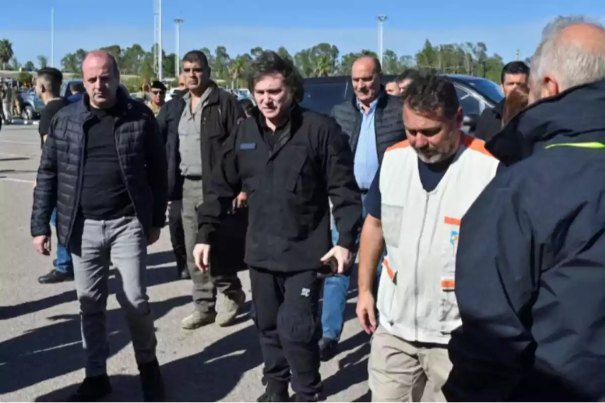 Un grupo de personas camina al aire libre en un área pavimentada con árboles y vehículos al fondo.