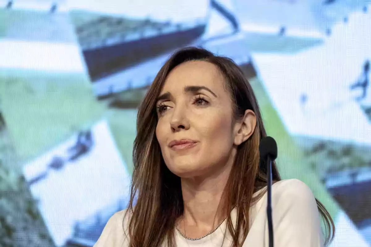 A person with brown hair and a pensive expression in front of a microphone, with a blurred background of light colors.