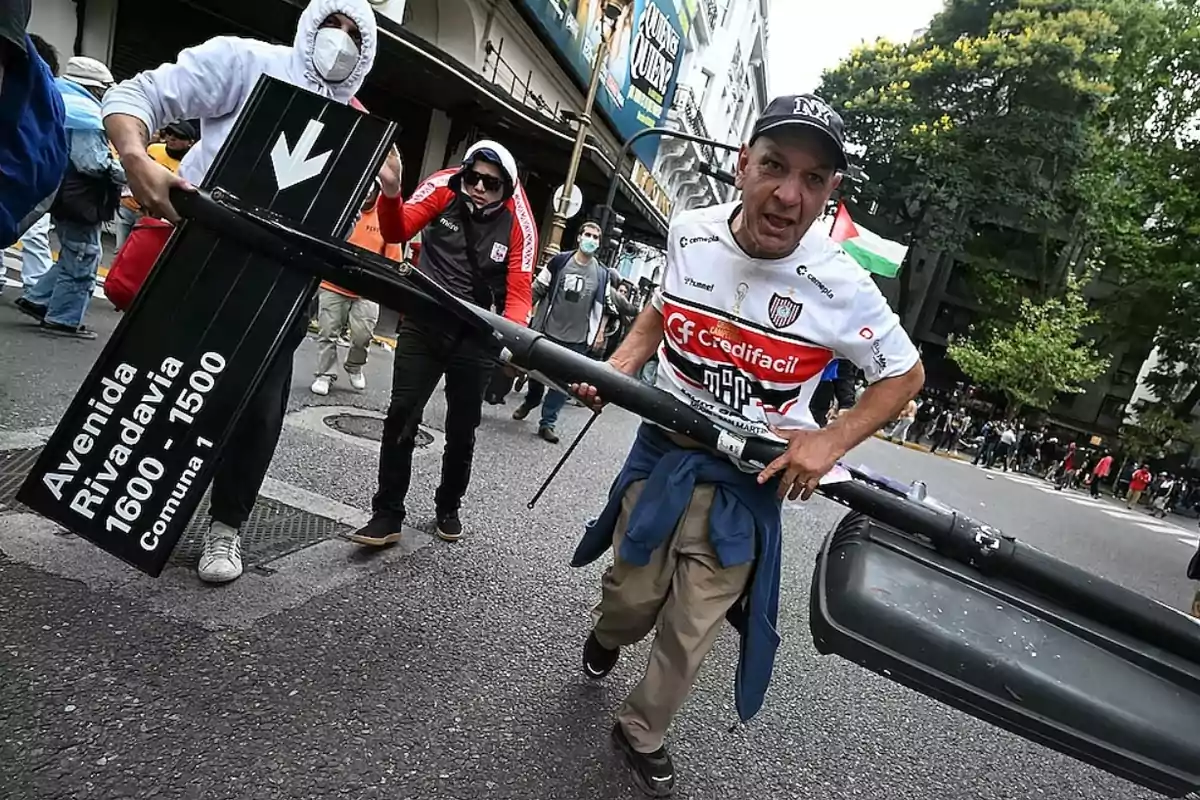 Un grupo de personas en una calle urbana, una de ellas lleva un poste de señalización que indica 