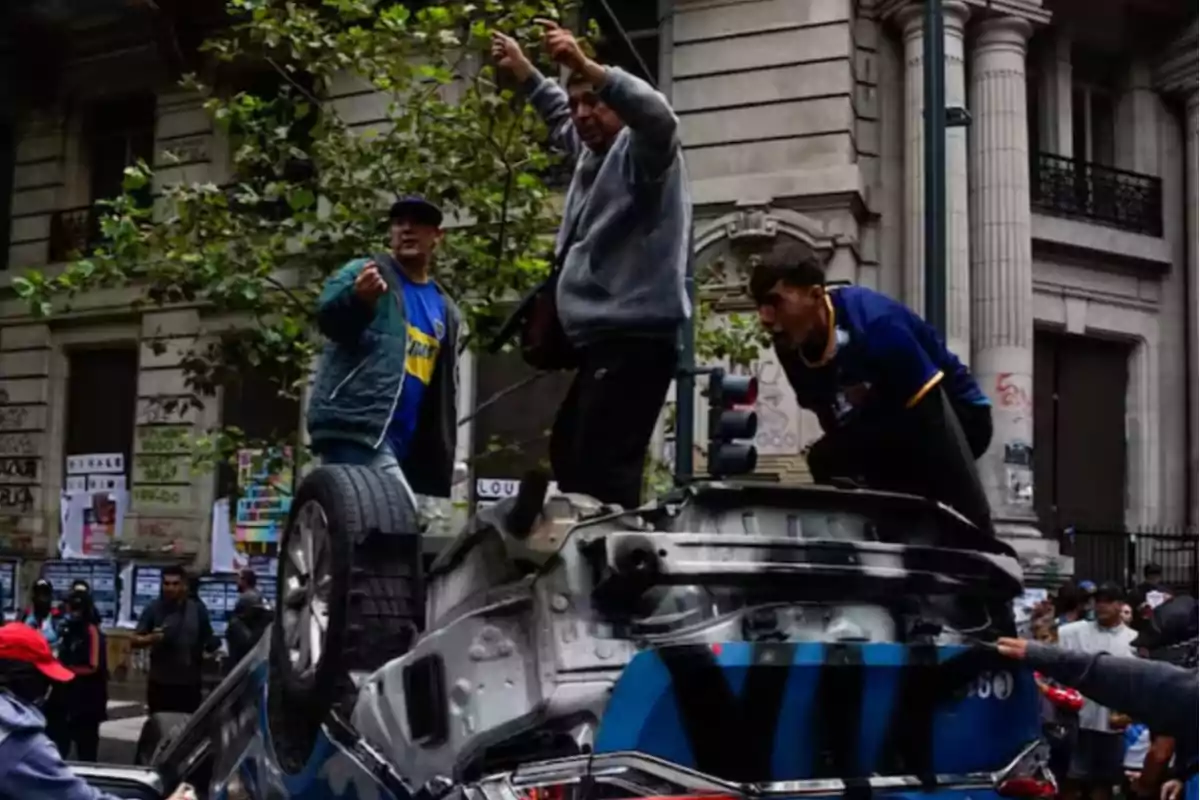 Un grupo de personas está de pie sobre un vehículo volcado en una calle urbana con edificios de fondo.