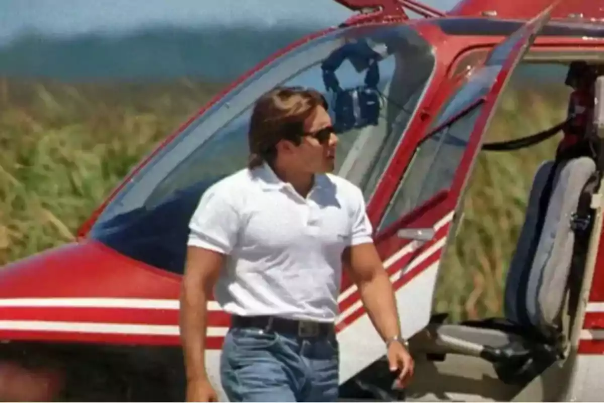 Un hombre con gafas de sol y camiseta blanca está de pie junto a un helicóptero rojo en un entorno al aire libre.