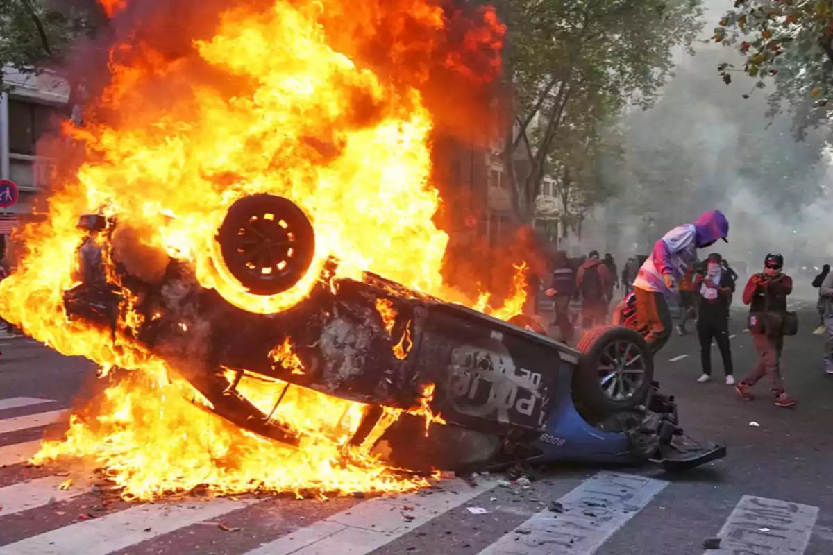 An overturned car in flames in the middle of a street with people around.