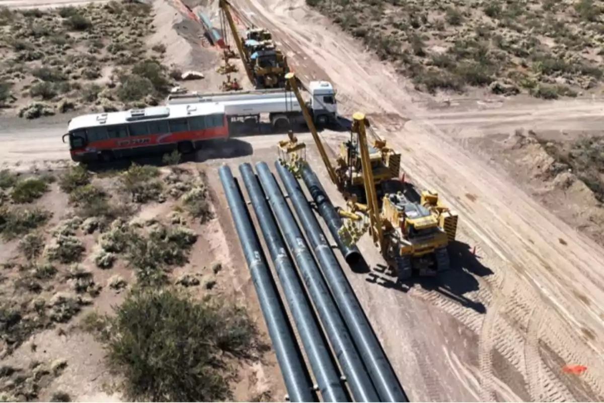 An aerial image shows heavy machinery and large pipes at a construction site in a desert area with a bus and a tanker truck on a dirt road.