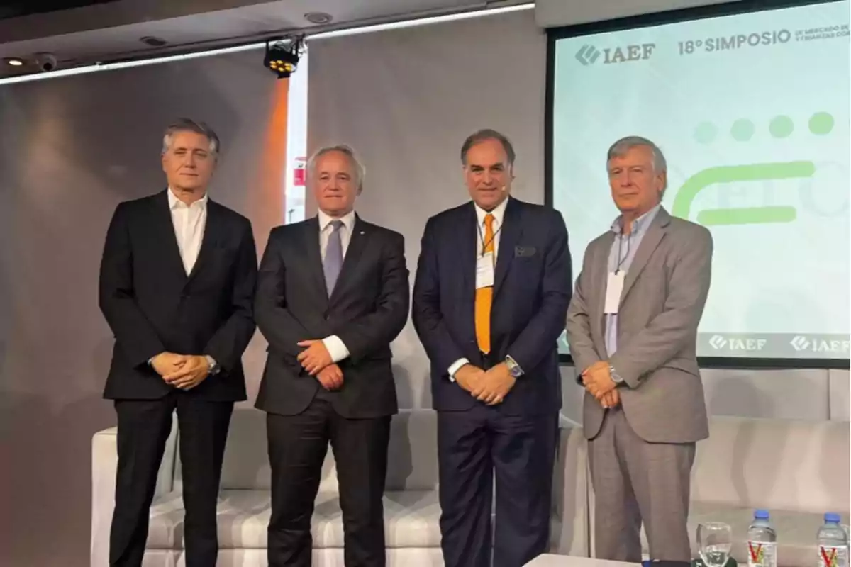 Four men standing on a stage with a background projector displaying the IAEF logo and the text "18th Symposium."