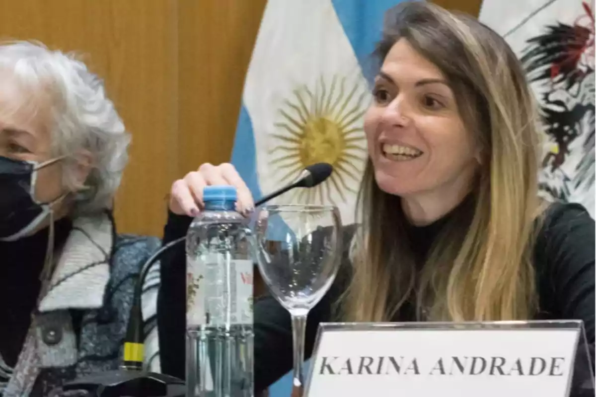 A smiling woman speaking into a microphone during an event with flags in the background.