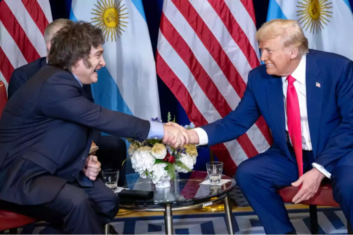 Two people shaking hands at a formal meeting with the flags of Argentina and the United States in the background.