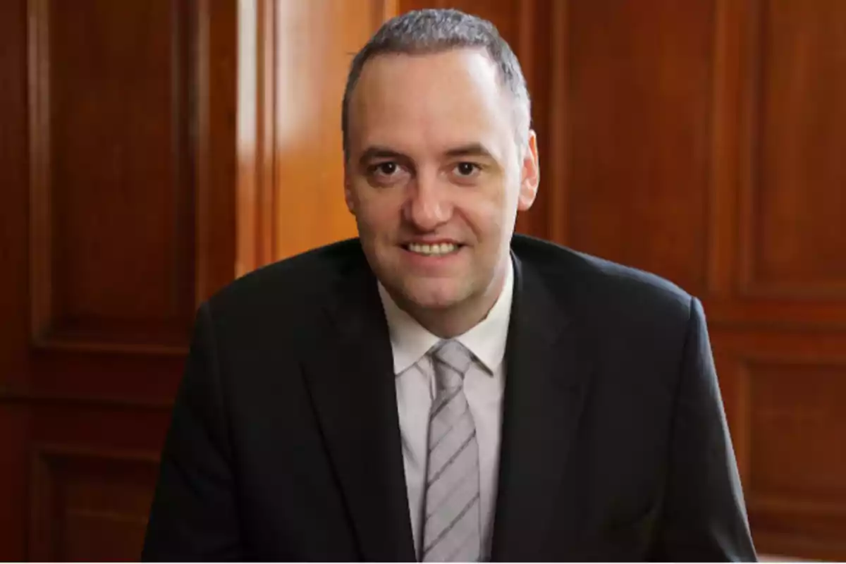 Un hombre con traje oscuro y corbata gris sonriendo frente a un fondo de madera. Un hombre con traje oscuro y corbata gris sonriendo frente a un fondo de madera.