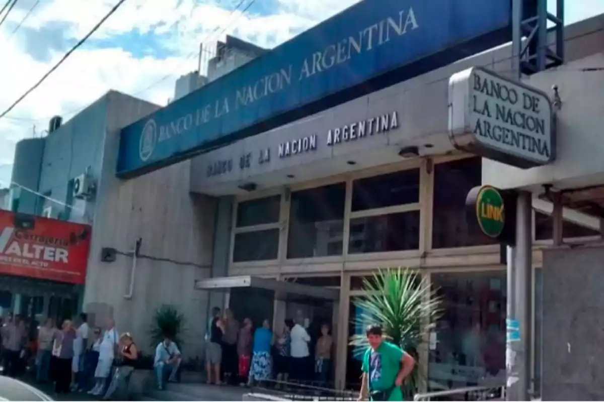 Personas haciendo fila afuera de una sucursal del Banco de la Nación Argentina en un día soleado.