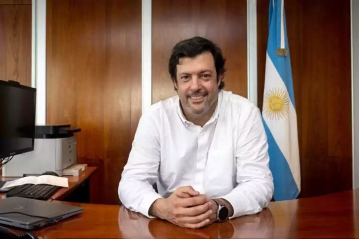 Un hombre con camisa blanca sonríe mientras está sentado en un escritorio de madera con una bandera de Argentina al fondo. Un hombre con camisa blanca sonríe mientras está sentado en un escritorio de madera con una bandera de Argentina al fondo.