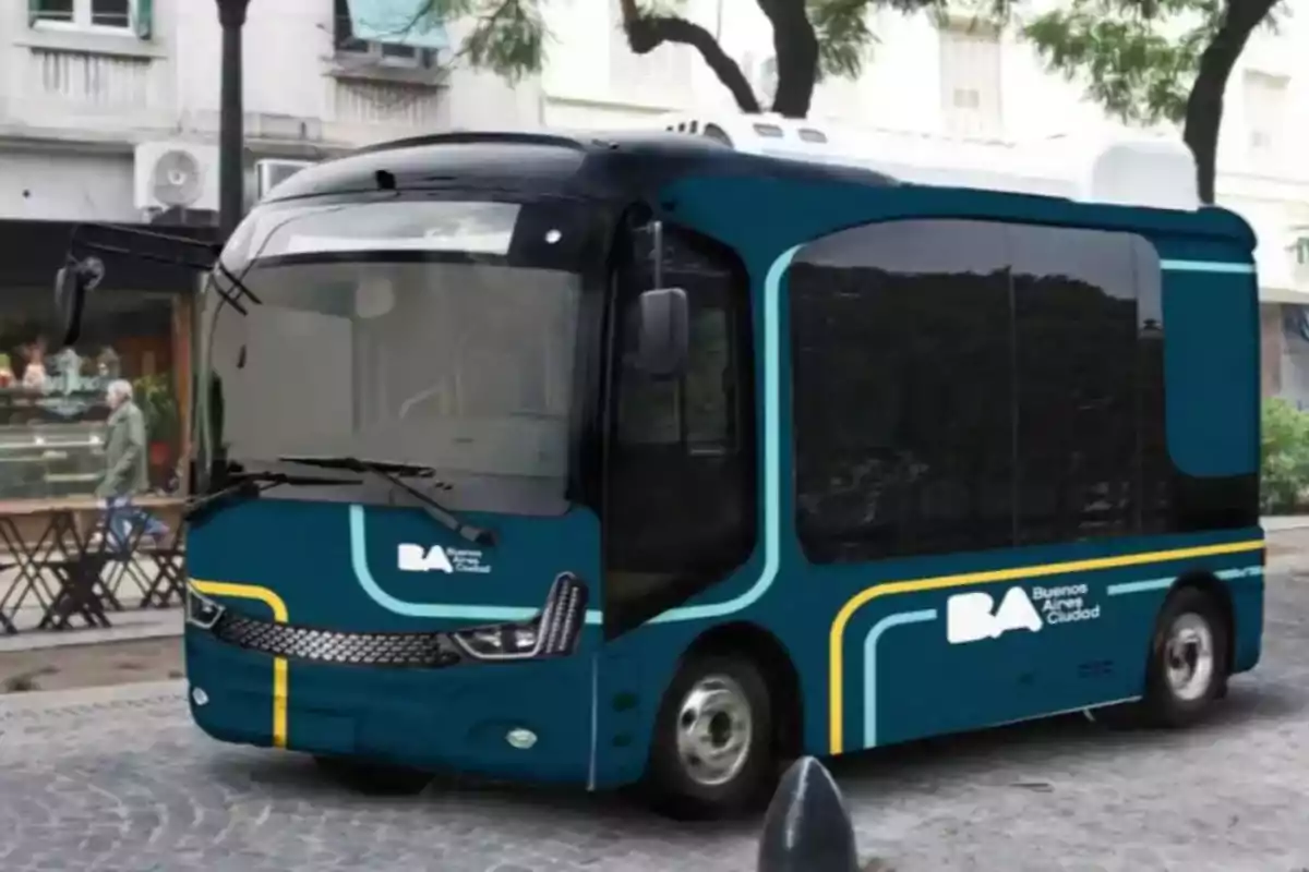 Un pequeño autobús eléctrico azul con detalles amarillos y blancos circula por una calle adoquinada en una ciudad, con árboles y edificios de fondo.
