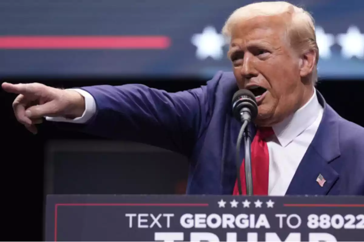 A man in a blue suit and red tie speaks into a microphone while pointing with his finger.