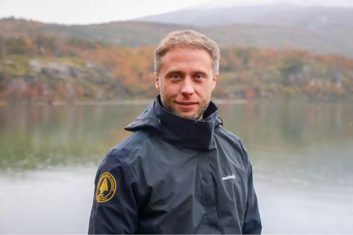 Un hombre con chaqueta oscura de pie frente a un lago con un paisaje otoñal de fondo. Un hombre con chaqueta oscura de pie frente a un lago con un paisaje otoñal de fondo.