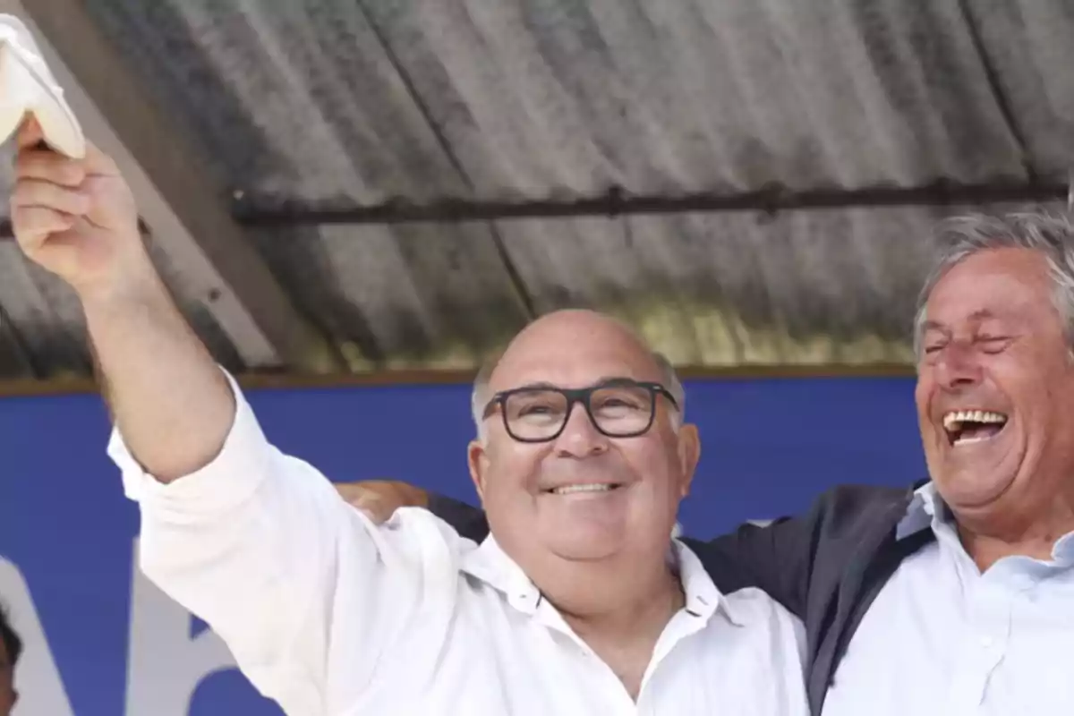 Dos hombres sonrientes, uno de ellos levantando un brazo, bajo un techo de metal.