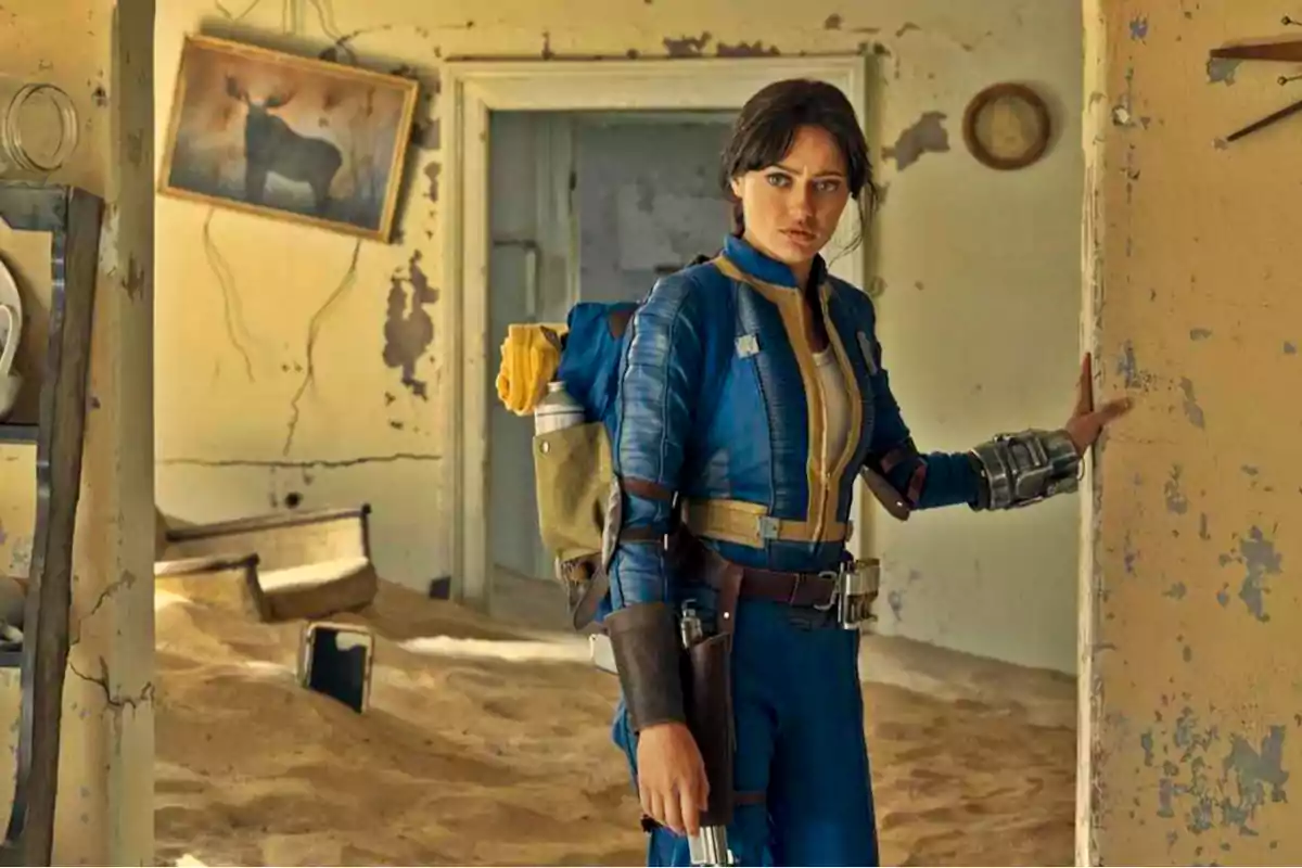Una persona con un traje azul y amarillo, similar al de un videojuego postapocalíptico, está de pie en una habitación deteriorada con arena en el suelo y paredes descascaradas, sosteniendo una pistola y llevando una mochila.