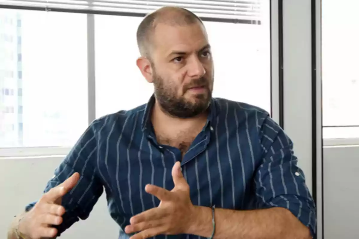 Un hombre con barba y camisa de rayas azules gesticula mientras habla en una oficina iluminada por la luz natural.
