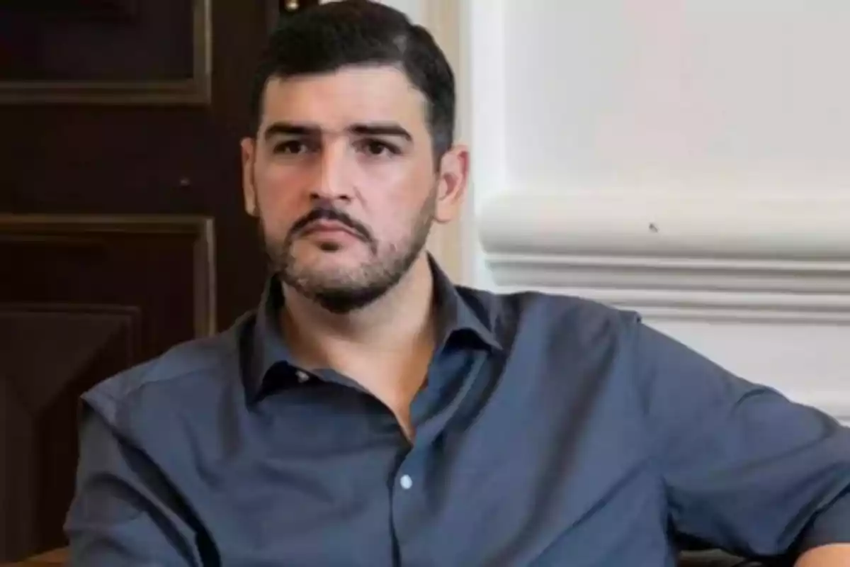 Un hombre con barba y camisa oscura está sentado en una habitación con paredes blancas y una puerta de madera. Un hombre con barba y camisa oscura está sentado en una habitación con paredes blancas y una puerta de madera.