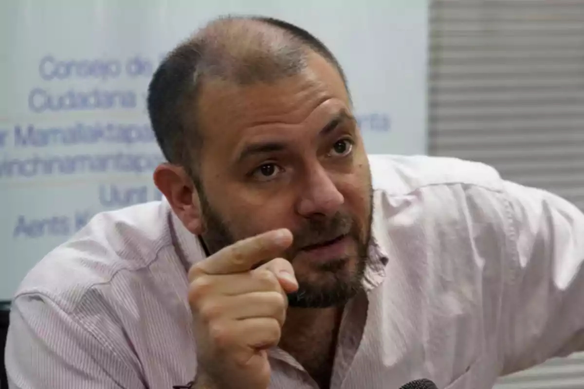 Un hombre con barba y camisa clara gesticula mientras señala con el dedo.