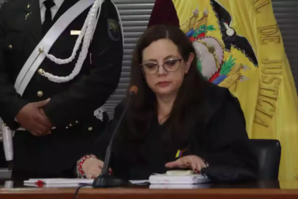 Una mujer sentada en una mesa con un micrófono frente a ella, junto a un hombre de uniforme militar, con una bandera detrás. Una mujer sentada en una mesa con un micrófono frente a ella, junto a un hombre de uniforme militar, con una bandera detrás.