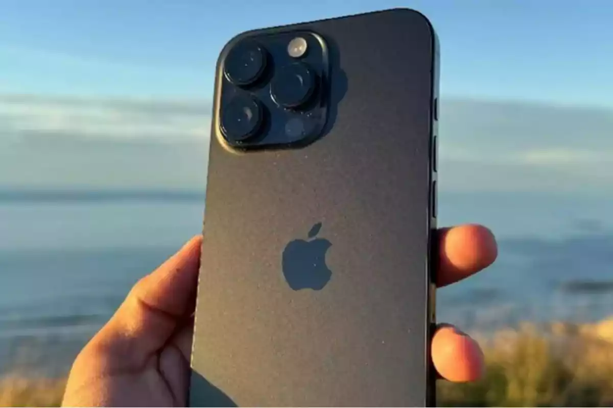 A hand holds a smartphone with a triple camera in an outdoor setting with the sea in the background. A hand holds a smartphone with a triple camera in an outdoor setting with the sea in the background.