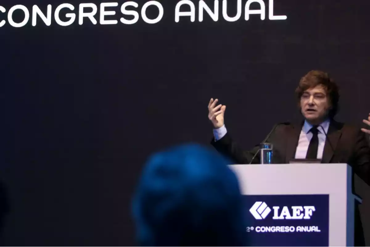 A man speaking at a podium during an annual conference, with a dark background and text that says "ANNUAL CONFERENCE."