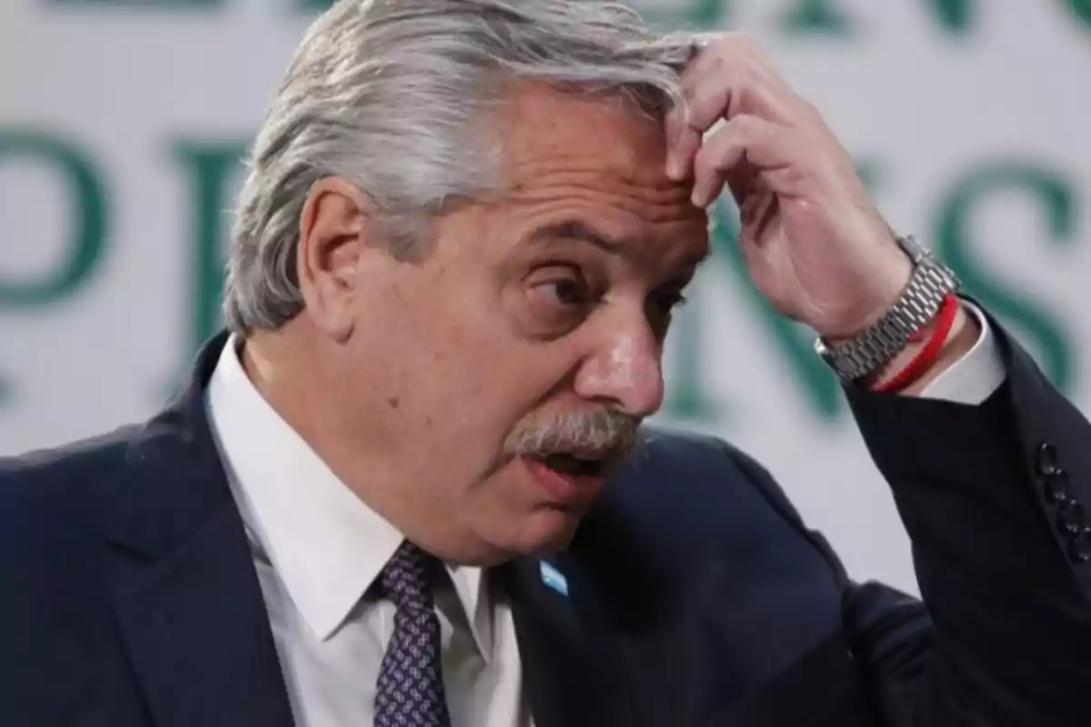 Un hombre de cabello canoso y bigote se toca la cabeza con expresión pensativa mientras viste un traje oscuro y corbata. Un hombre de cabello canoso y bigote se toca la cabeza con expresión pensativa mientras viste un traje oscuro y corbata.