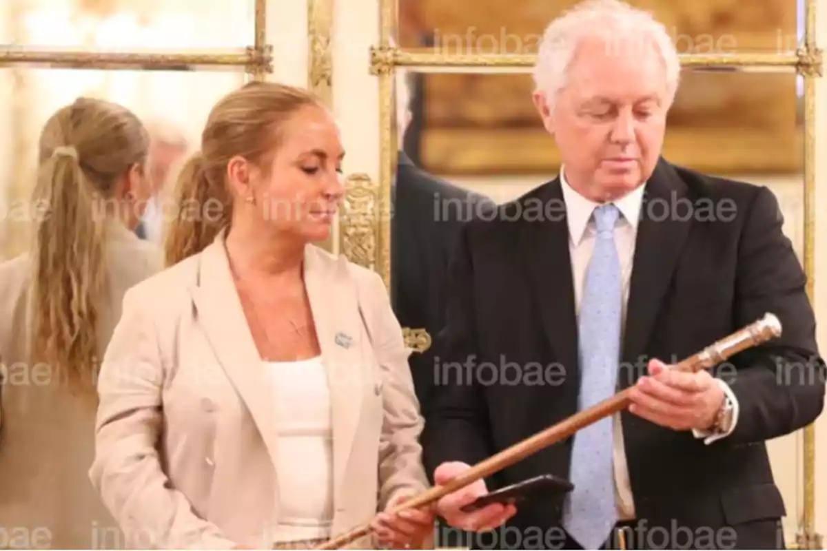 Dos personas en un entorno formal, una de ellas sostiene un bastón ceremonial mientras la otra observa. Dos personas en un entorno formal, una de ellas sostiene un bastón ceremonial mientras la otra observa.