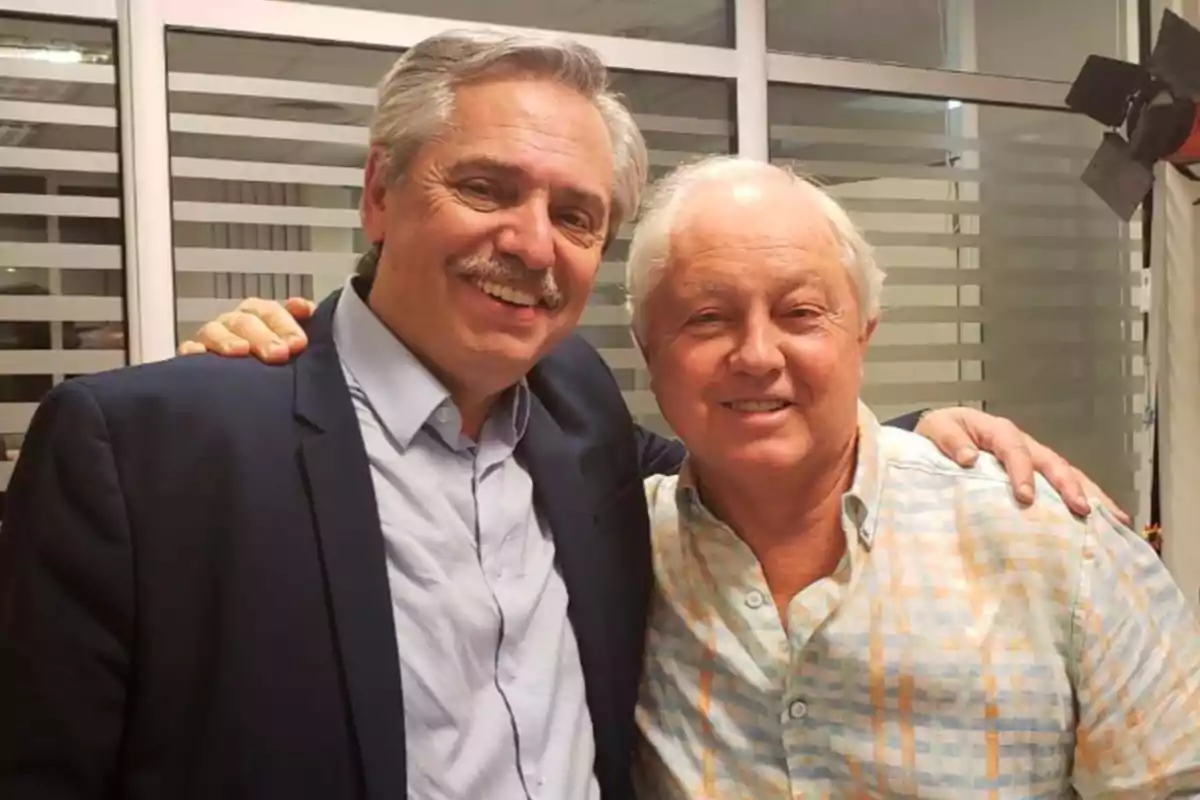 Dos hombres sonrientes posan juntos en una oficina con ventanas de fondo.