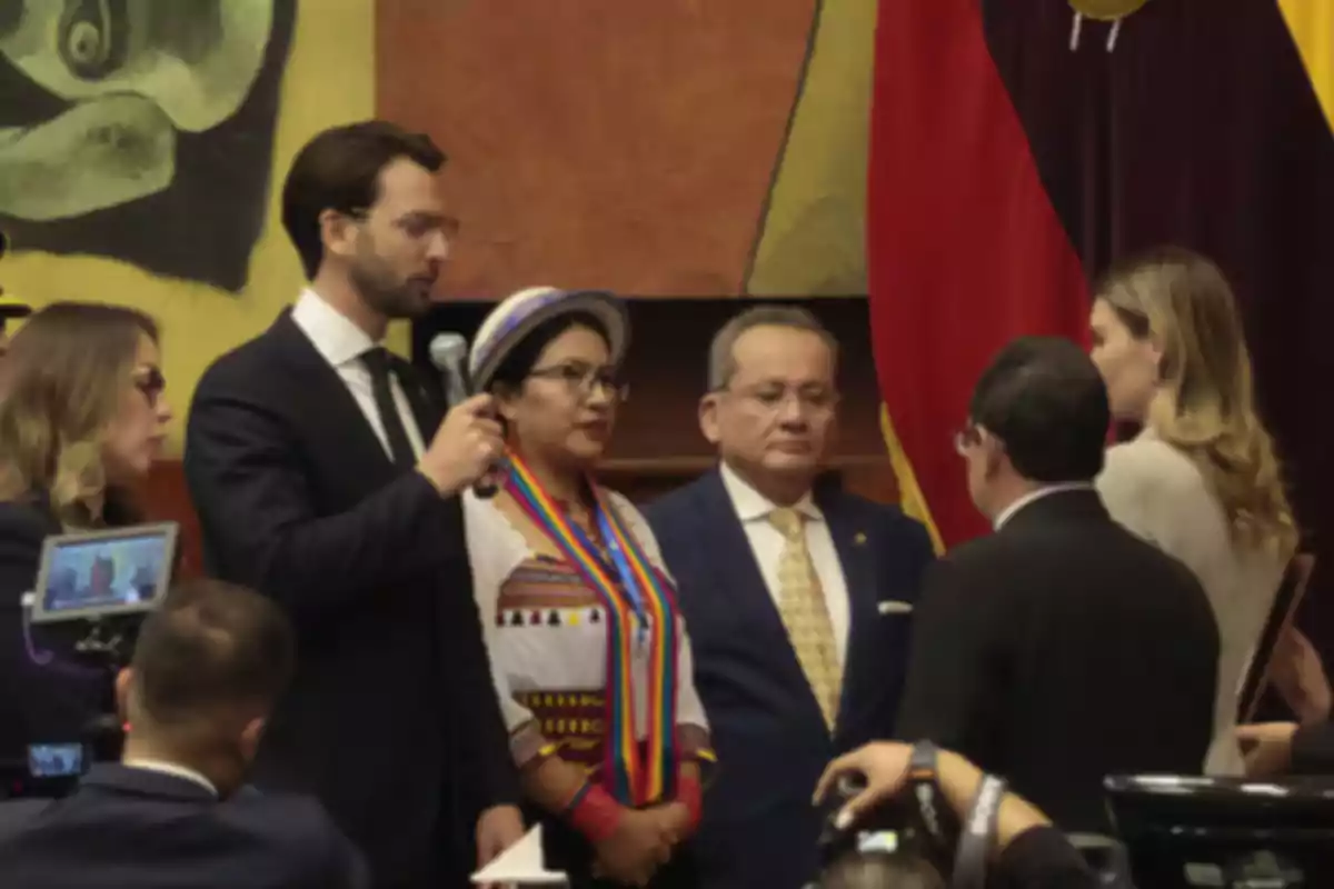 Un grupo de personas en un evento formal, algunas de ellas sostienen micrófonos y cámaras, con banderas y arte en el fondo. Un grupo de personas en un evento formal, algunas de ellas sostienen micrófonos y cámaras, con banderas y arte en el fondo.
