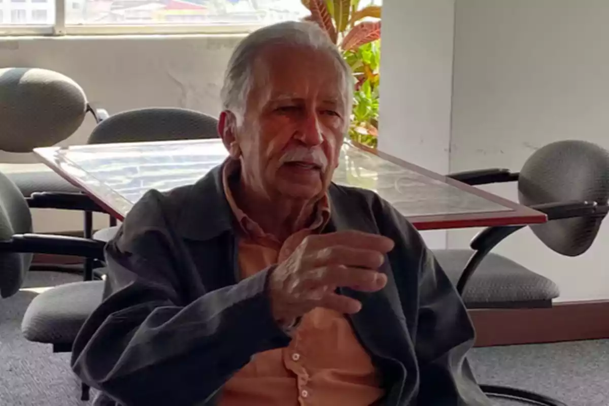 An older man with a mustache and gray hair is sitting in an office, gesturing as he speaks. An older man with a mustache and gray hair is sitting in an office, gesturing as he speaks.