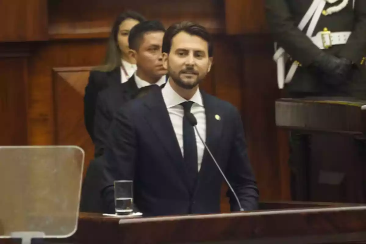 Un hombre de traje oscuro habla en un podio en un entorno formal con personas de pie detrás de él. Un hombre de traje oscuro habla en un podio en un entorno formal con personas de pie detrás de él.