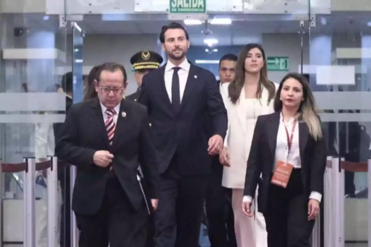Un grupo de personas vestidas formalmente camina por un pasillo con puertas de vidrio y un letrero de salida de emergencia arriba.