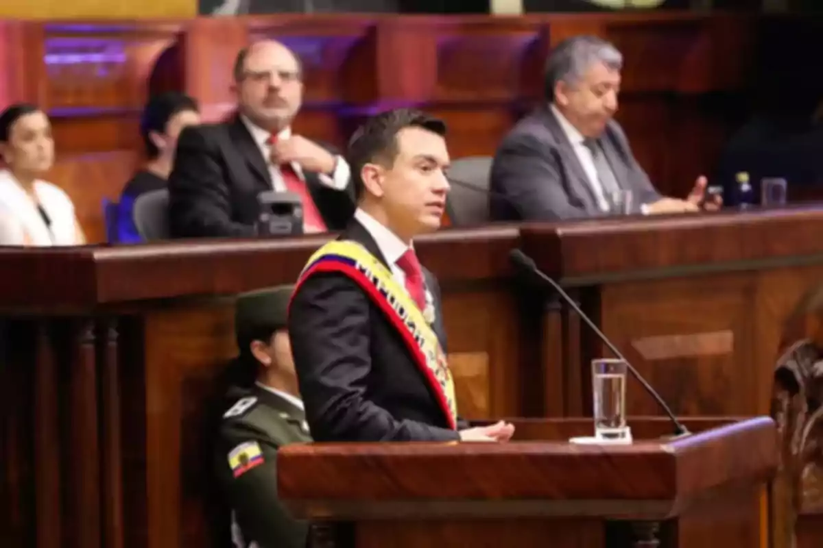 A man with a presidential sash speaks at a podium in a formal setting with several people seated behind him.