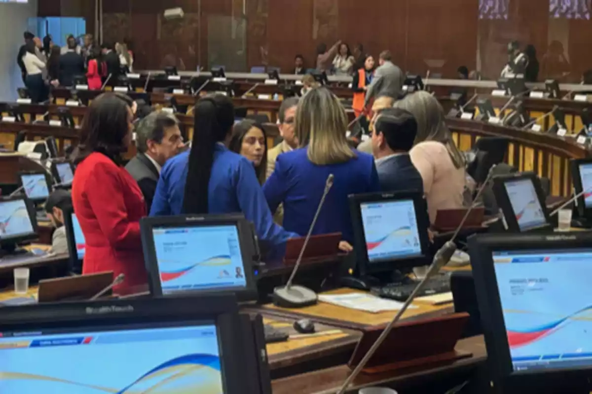 A group of people gathers in the center of a conference room or parliament, surrounded by desks with monitors and microphones. A group of people gathers in the center of a conference room or parliament, surrounded by desks with monitors and microphones.