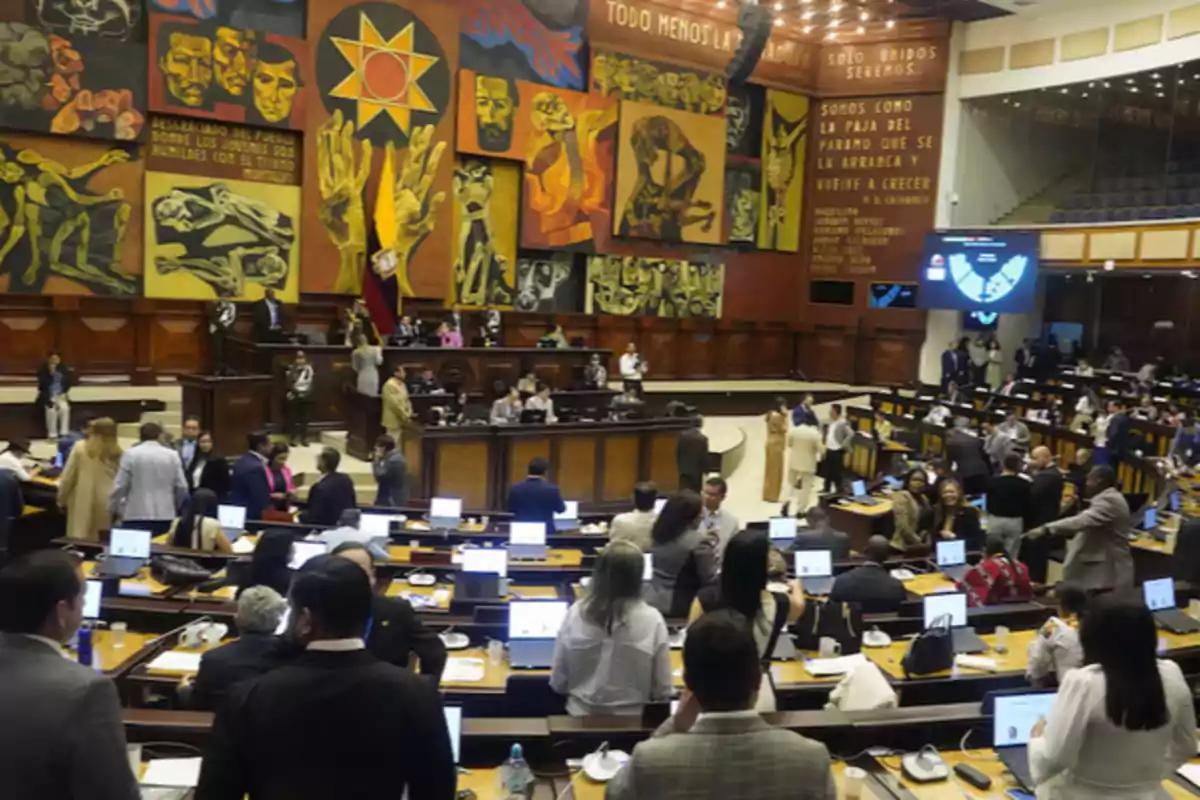 Una asamblea legislativa con personas reunidas en un salón amplio, decorado con murales coloridos y una bandera nacional. Una asamblea legislativa con personas reunidas en un salón amplio, decorado con murales coloridos y una bandera nacional.