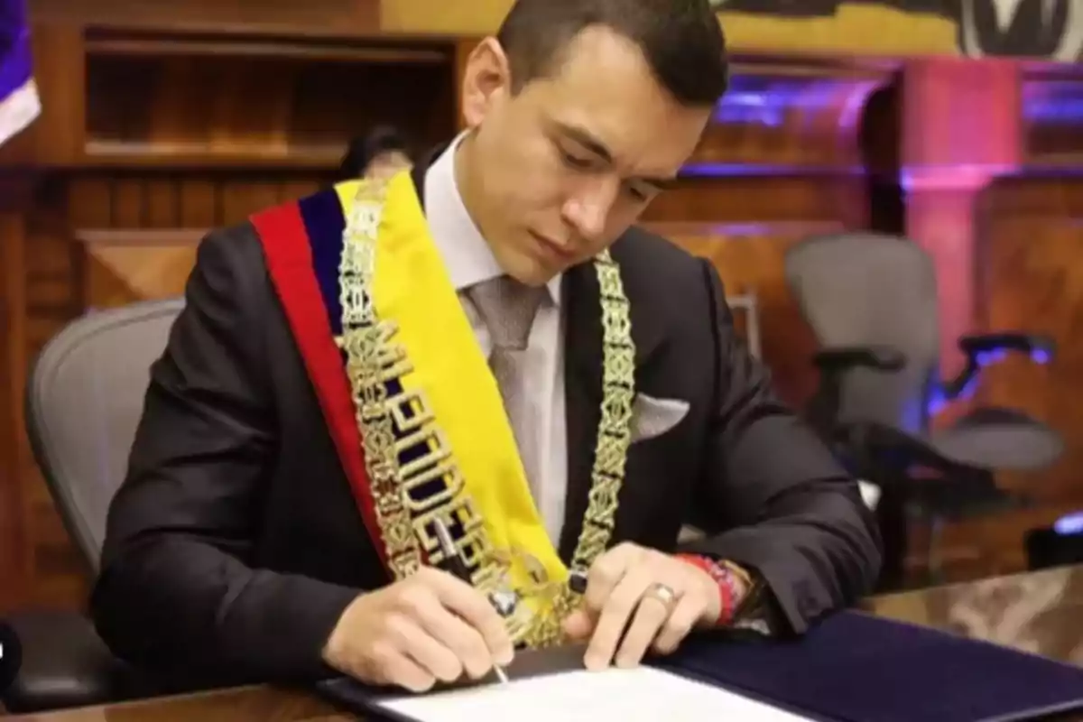 Un hombre con traje formal y banda presidencial firma un documento en un escritorio.