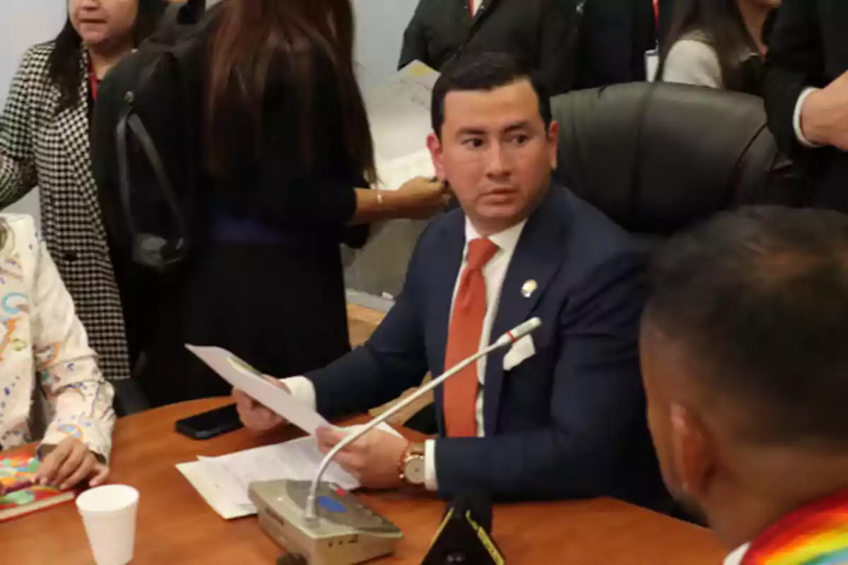 A man in a suit and orange tie is sitting at a table, holding papers and looking to the side, surrounded by other people in an office or meeting setting.