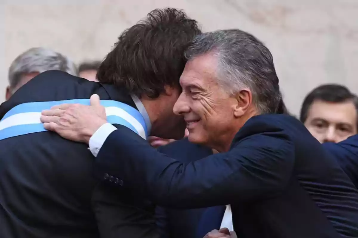 Dos personas se abrazan en un evento formal, una de ellas lleva una banda celeste y blanca. Dos personas se abrazan en un evento formal, una de ellas lleva una banda celeste y blanca.