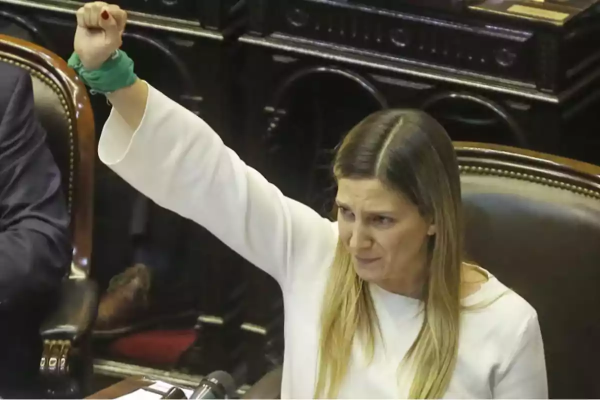 Una mujer con el puño levantado y un pañuelo verde en la muñeca, sentada en un entorno formal. Una mujer con el puño levantado y un pañuelo verde en la muñeca, sentada en un entorno formal.