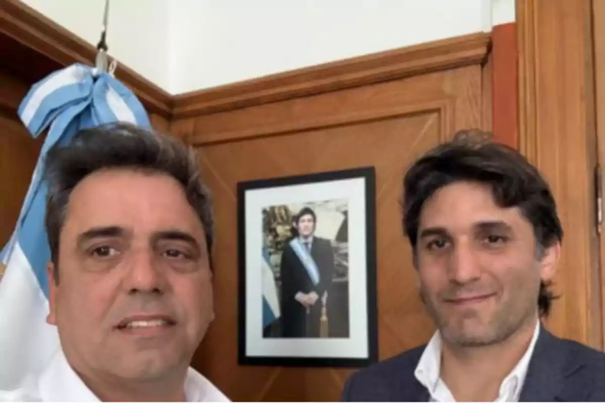 Two people pose for a photo in front of a framed portrait of a man with a presidential sash, with an Argentine flag in the background.