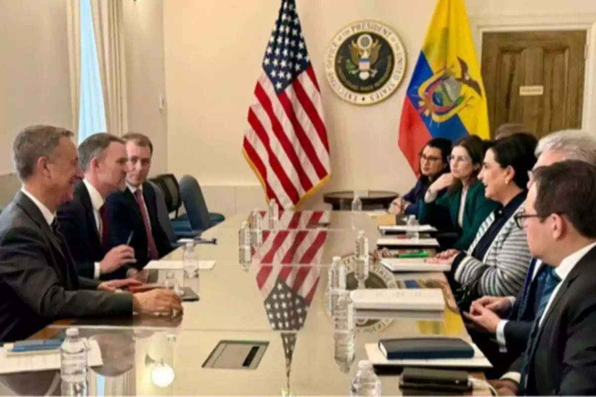 Un grupo de personas en una reunión formal sentadas a ambos lados de una mesa con banderas de Estados Unidos y Ecuador en el fondo. Un grupo de personas en una reunión formal sentadas a ambos lados de una mesa con banderas de Estados Unidos y Ecuador en el fondo.