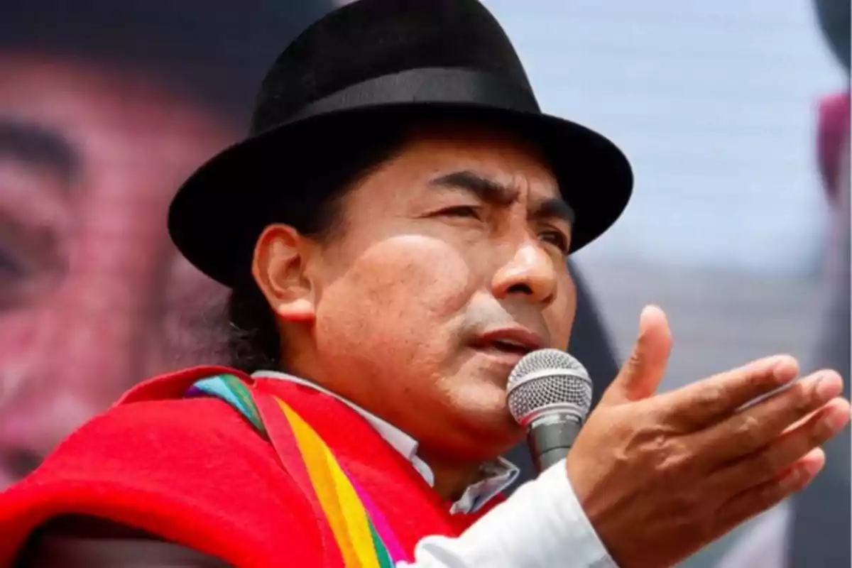Un hombre con sombrero y poncho rojo habla al micrófono en un evento al aire libre. Un hombre con sombrero y poncho rojo habla al micrófono en un evento al aire libre.