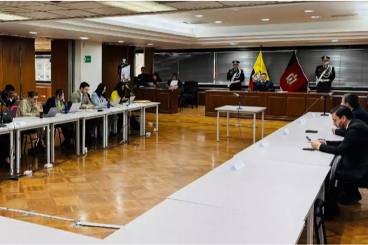 Una sala de reuniones con varias personas sentadas en mesas largas, algunas usando computadoras portátiles, mientras al fondo hay dos banderas y personal uniformado.