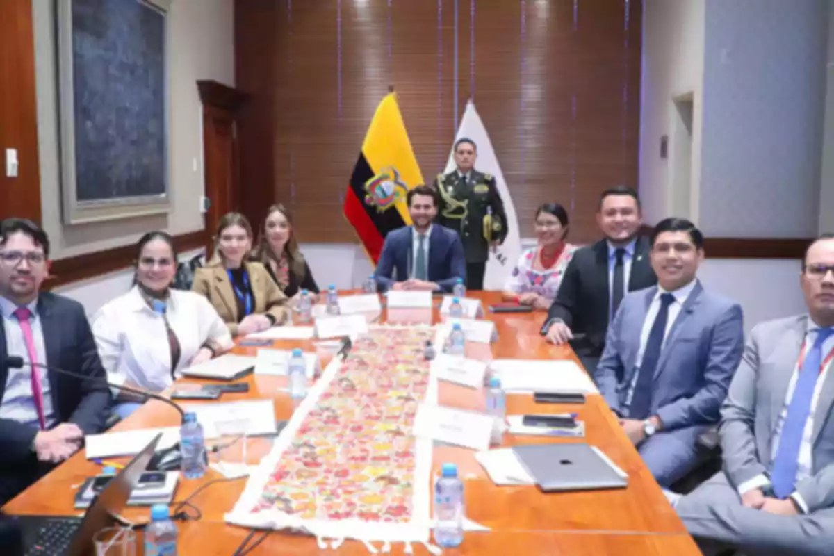 A group of people in a formal meeting sitting around a table with Ecuador flags in the background.