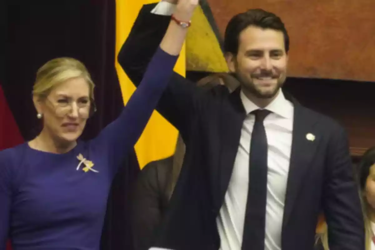 Two people standing, one raising the other's arm, both smiling and dressed formally, with a background of flags and curtains. Two people standing, one raising the other's arm, both smiling and dressed formally, with a background of flags and curtains.